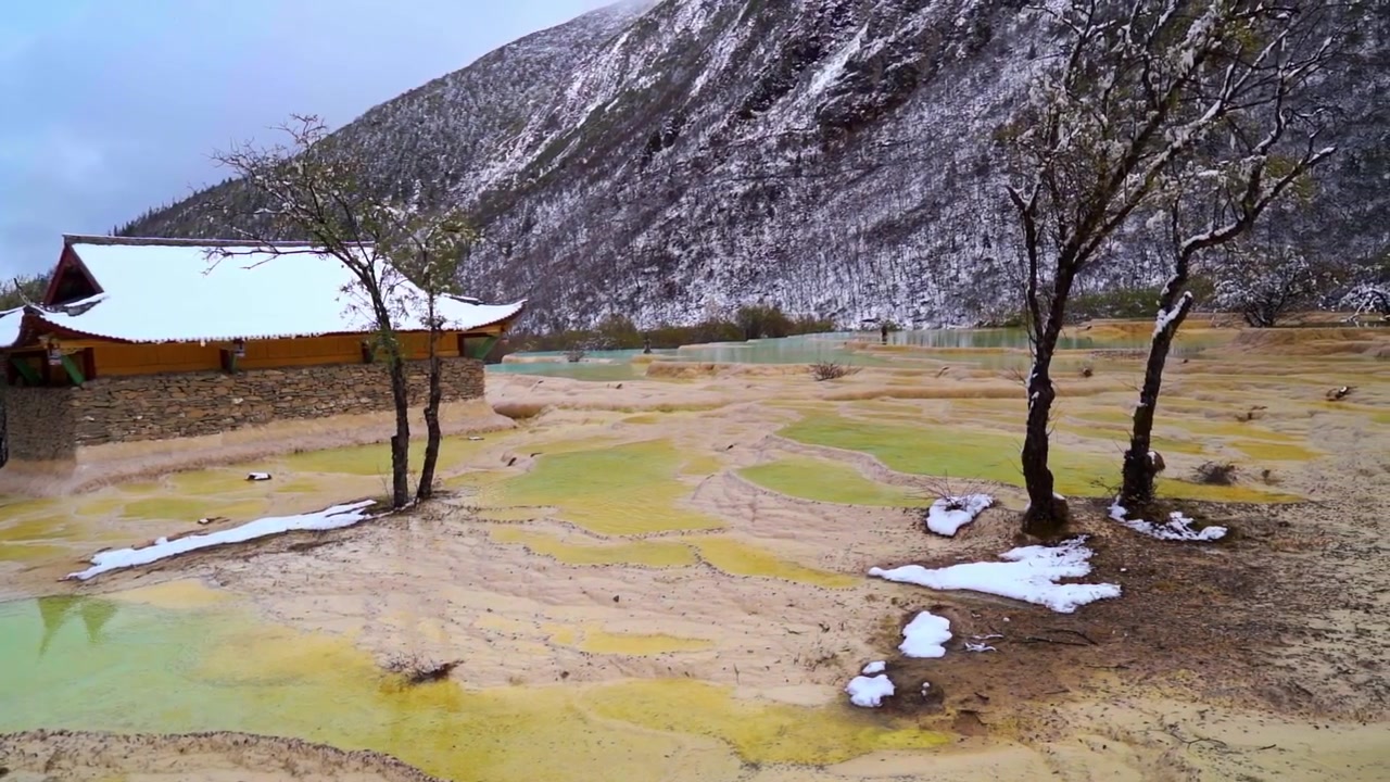 Karla kaplı yamaçların altında, geleneksel taş duvarlı bir köşkün yanında yer alan altın sarısı mineral terasların ve açık yeşil havuzların geniş, yavaş kaydırma çekimi.