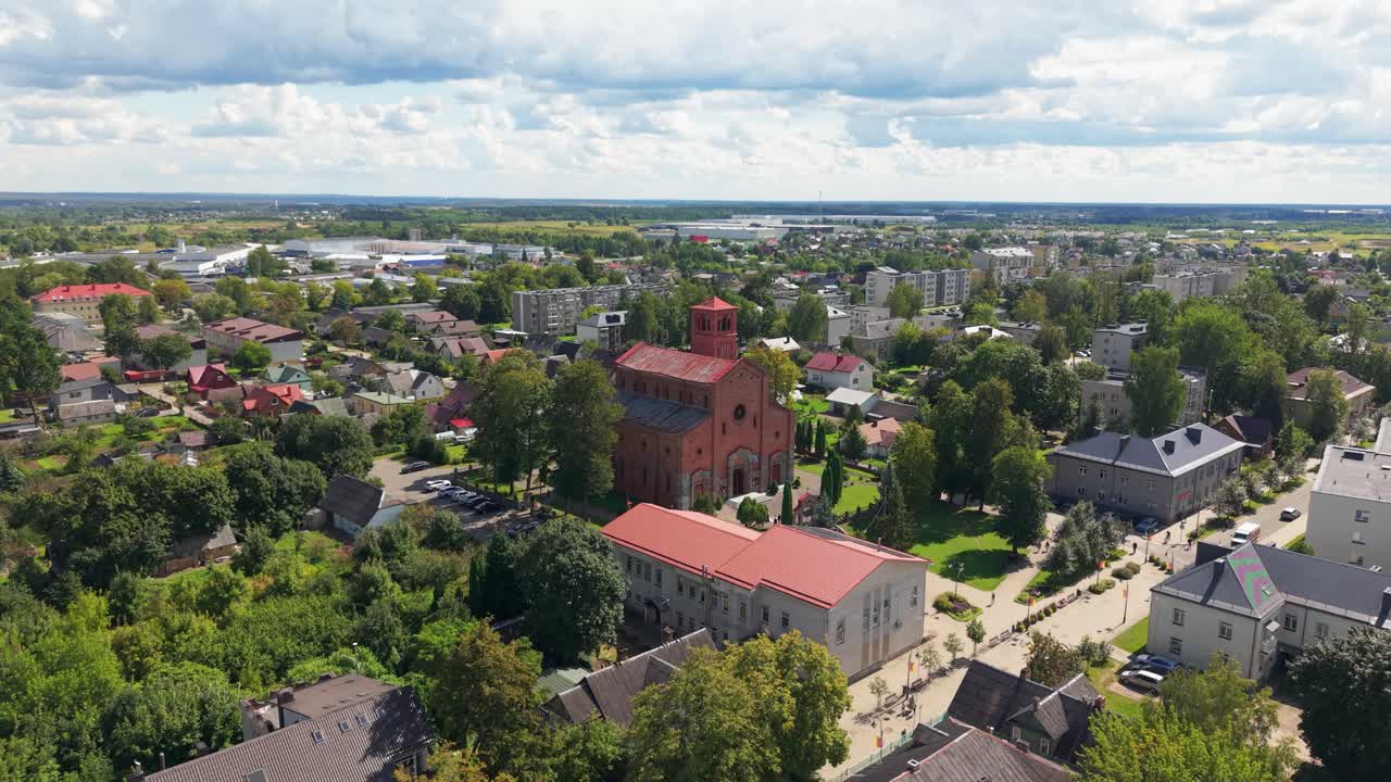 Aerial view of Lentvaris town in Lithuania showcasing its central red brick church surrounded by trees, homes, and modern residential areas under a partly cloudy summer sky