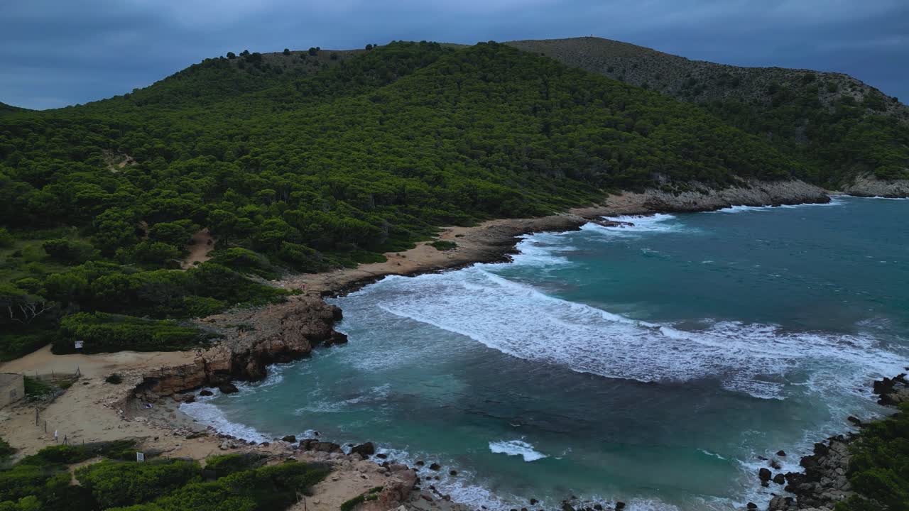 Capturing the varied geography of Cala Agulla in Majorca. It shows heavy surf hitting the sandy beach and rocky coves, contrasting the wild nature with nearby tourist development
