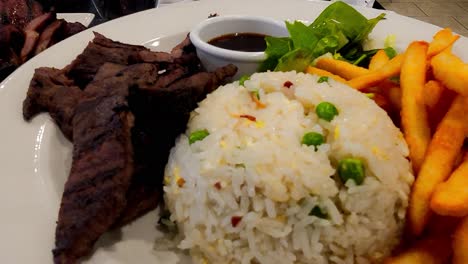 rotating-plate-of-Manila-Pilipino-food-grilled-beef-Baka-Pares-with-rice-mixed-with-peas-and-pieces-of-scrambled-eggs,-fries,-green-salad,-and-gravy-in-a-small-bowl