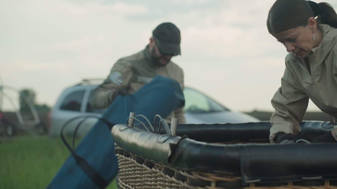 close up of woman and man removing ropes and carabiners from wicker balloon basket on grassy field under cloudy sky, focus on careful hand movements and safety check