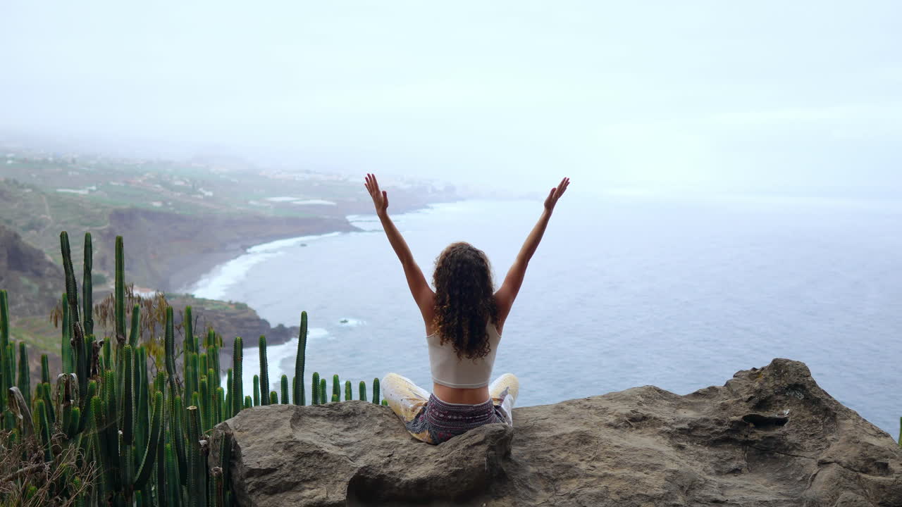 On an island's mountain, a young woman practices yoga, sitting on a rock, meditating in Lotus position, overlooking the ocean