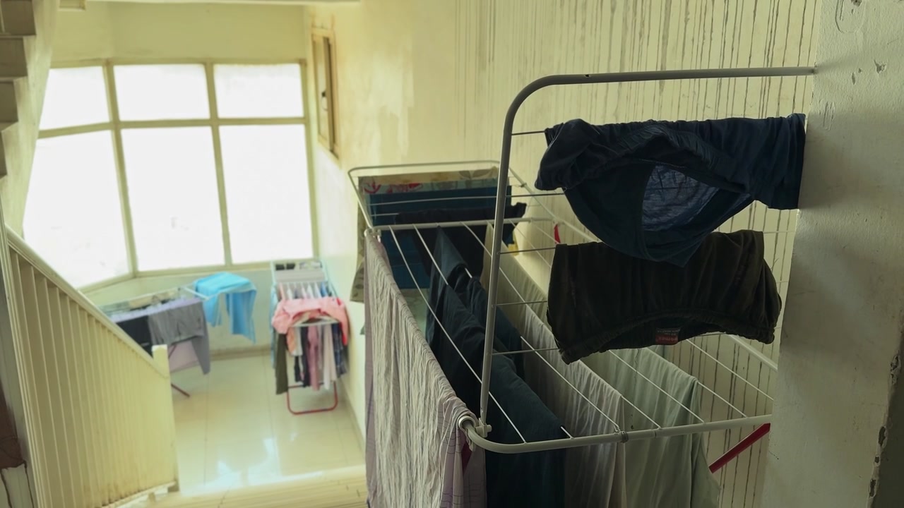 Washed clothes and laundry hang on metal drying racks in a bright residential apartment stairwell. Natural daylight streams through large windows onto the tiled landing of this indoor living space.