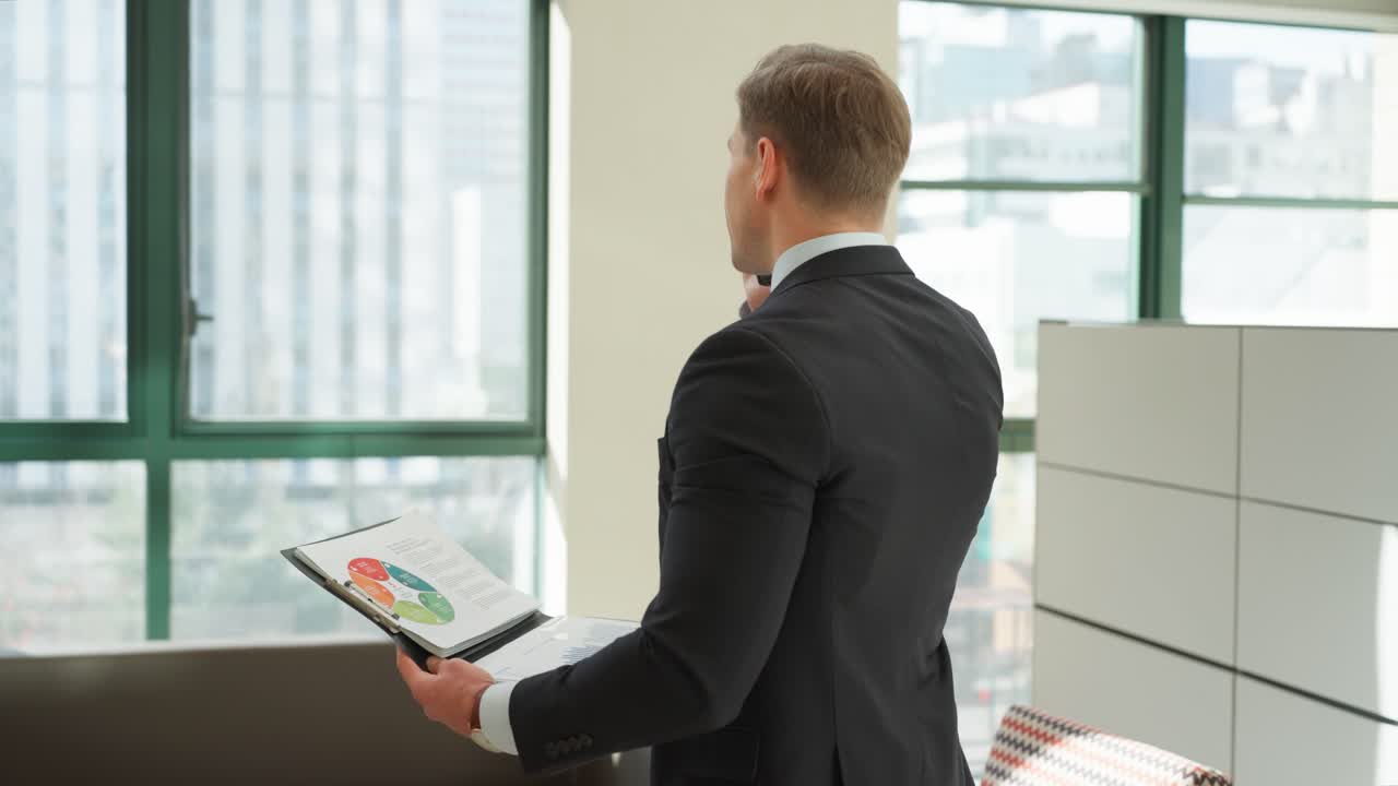 Un uomo caucasico felice sulla trentina, vestito in giacca e cravatta, parla al telefono mentre tiene in mano un rapporto dati o dei documenti in un ufficio luminoso - ripresa in parallasse
