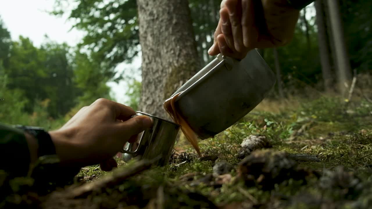 Een close-up, vanuit een lage hoek genomen shot van twee vrienden die een versgemaakte warme drank delen op hun kampeerplek in een prachtig, bemost bos