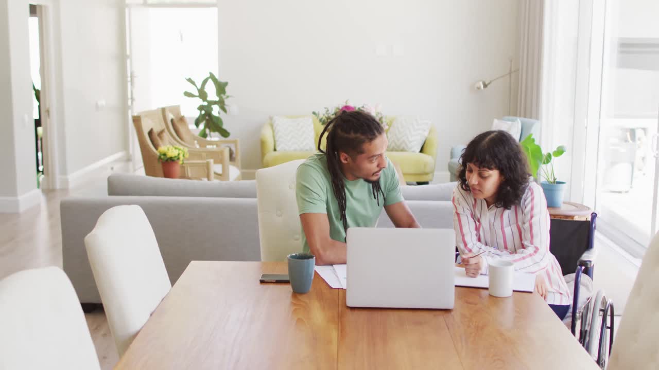 mujer biracial feliz en silla de ruedas y pareja masculina usando computadora portátil y hablando en la sala de estar
