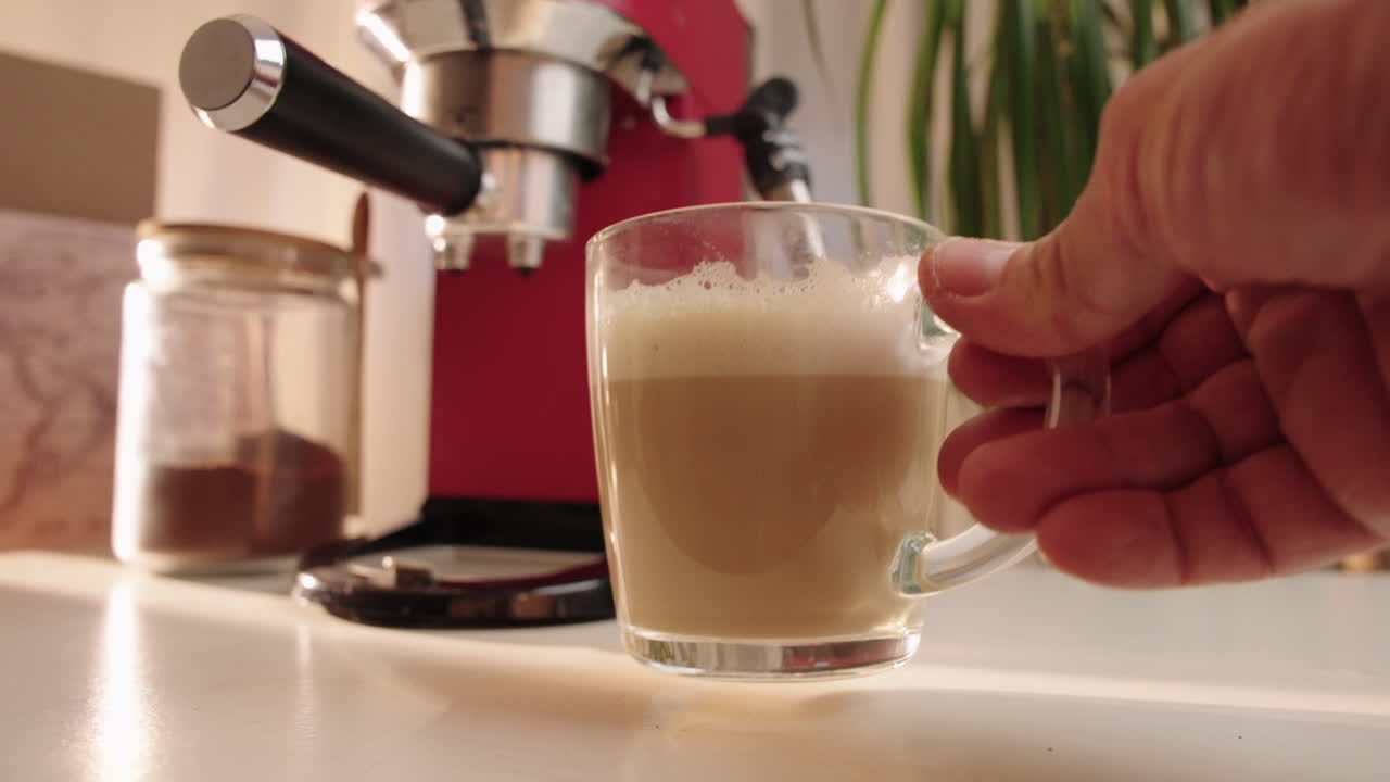 Espresso fresco in una tazza di vetro trasparente su un bancone della cucina. Movimento circolare della telecamera verso l'interno che evidenzia la texture della crema sotto una calda luce solare naturale, con macchina del caffè e pianta sullo sfondo