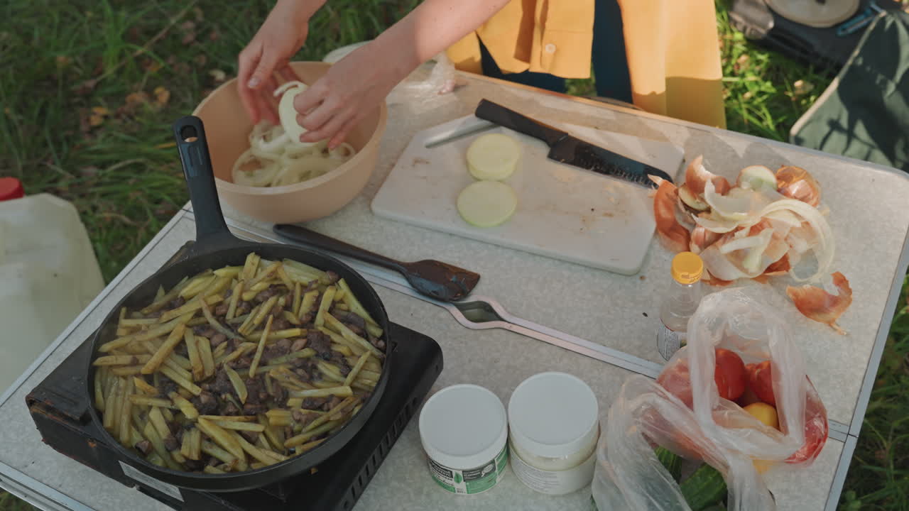 Mujer colocando cebollas en rodajas en un tazón junto a una sartén caliente con patatas fritas en una mesa de camping. Vista cenital que captura el movimiento de servir, productos frescos y utensilios, con una suave luz solar y un claro de césped como marco.