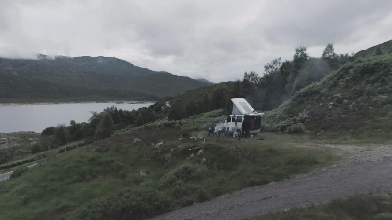 Close drone shot showing a campervan wild camping with BBQ smoke and Loch Beag and cloudy mountains in the background in Scottish Highlands