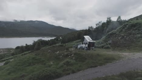 close drone shot showing a campervan wild camping with bbq smoke and loch beag and cloudy mountains in the background in scottish highlands