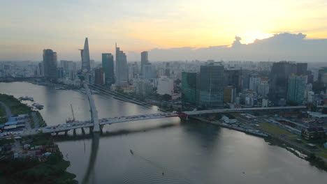 Flying-at-Sunset-over-Nearly-Constructed-Bridge-on-Saigon-River-in-Downtown-of-Ho-Chi-Minh-City-Vietnam,-Towers-and-Buildings-in-Background