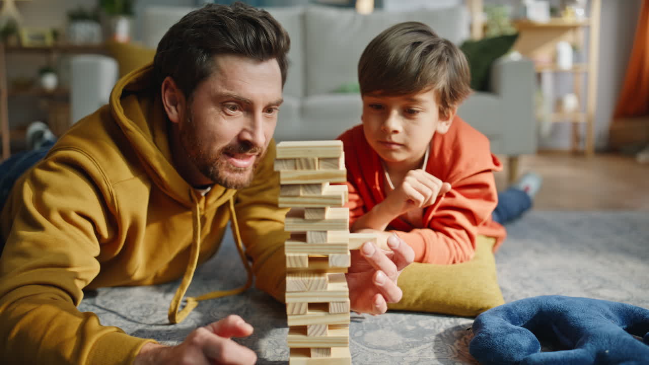 Padre e figlio che giocano a Jenga sdraiati sul pavimento, primo piano. Uomo sorridente e bambino giocoso che giocano.