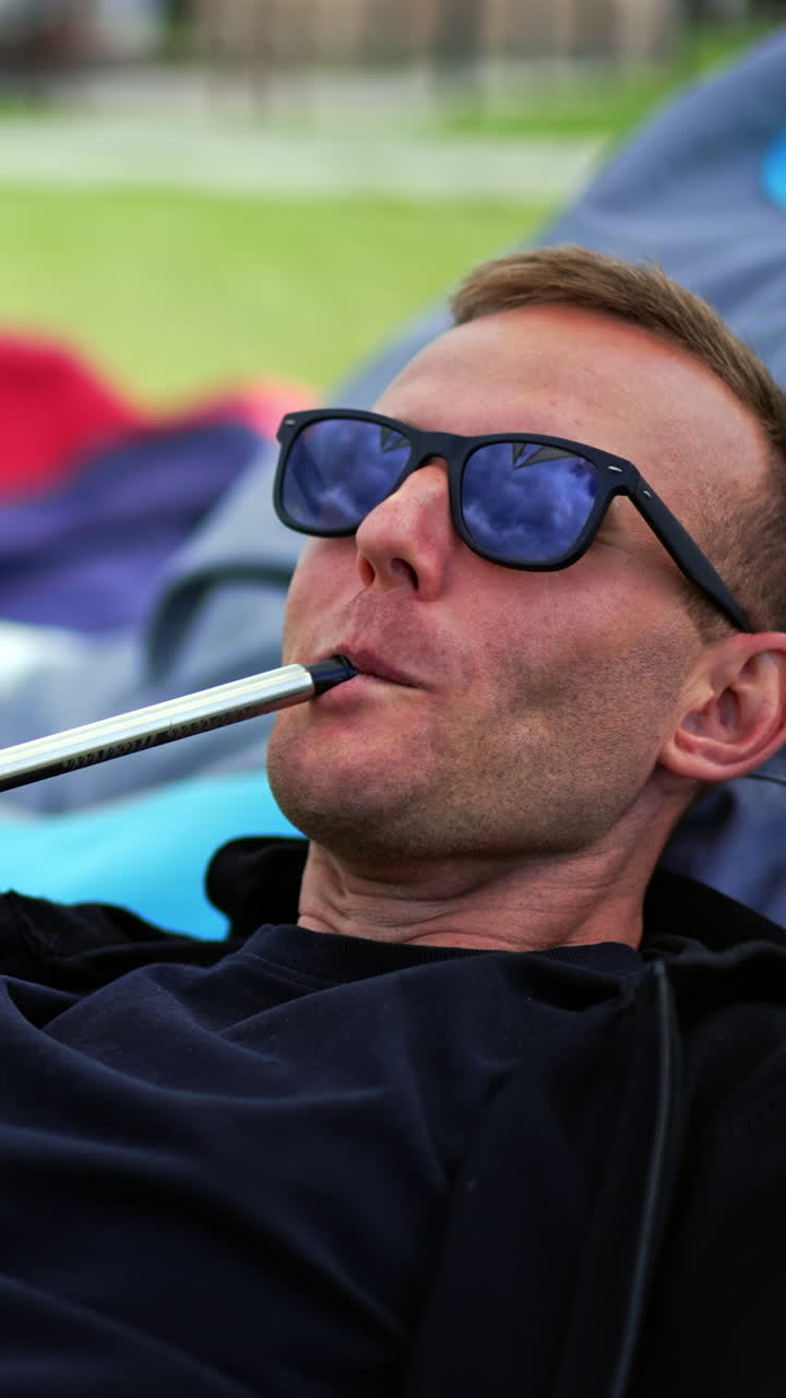 Smiling male freelancer working on computer combining it with rest outdoors. Man smoking hookah producing thick smoke. Vertical video