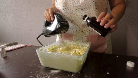 Mujer-Latina-Usando-Un-Delantal-Preparando-Cocinar-Horneando-Un-Pastel-Vertiendo-Extracto-De-Vainilla-En-La-Mezcla-De-Mantequilla-Y-Mezclando