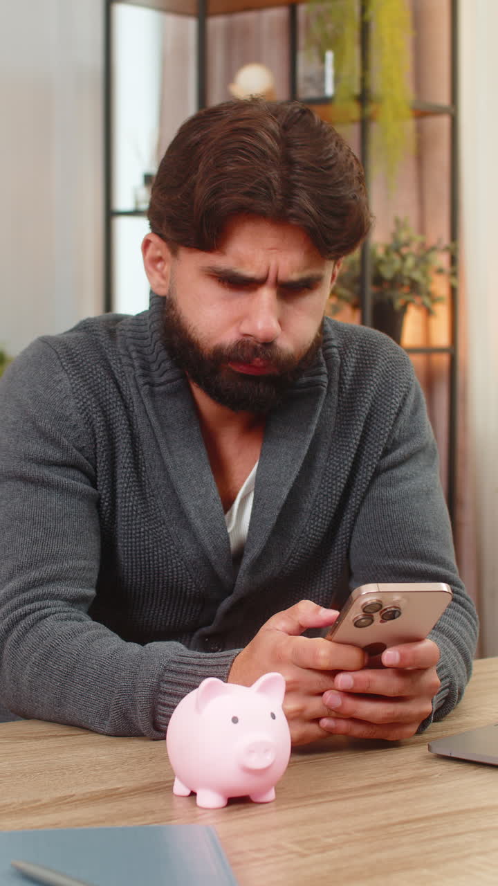Uomo indiano apre un salvadanaio vuoto, sospira e scrive un piano di budget mensile sullo smartphone per raggiungere i suoi obiettivi di risparmio