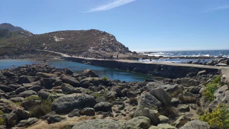 Gente-Caminando-Por-El-Paso-Elevado-Que-Cruza-El-Mar-Con-Las-Olas-Rompiendo-En-El-Fondo-En-Un-Día-Soleado-Sin-Nubes,-Tiro-Panorámico-Bloqueado,-Islas-Cíes,-Pontevedra,-Galicia,-España