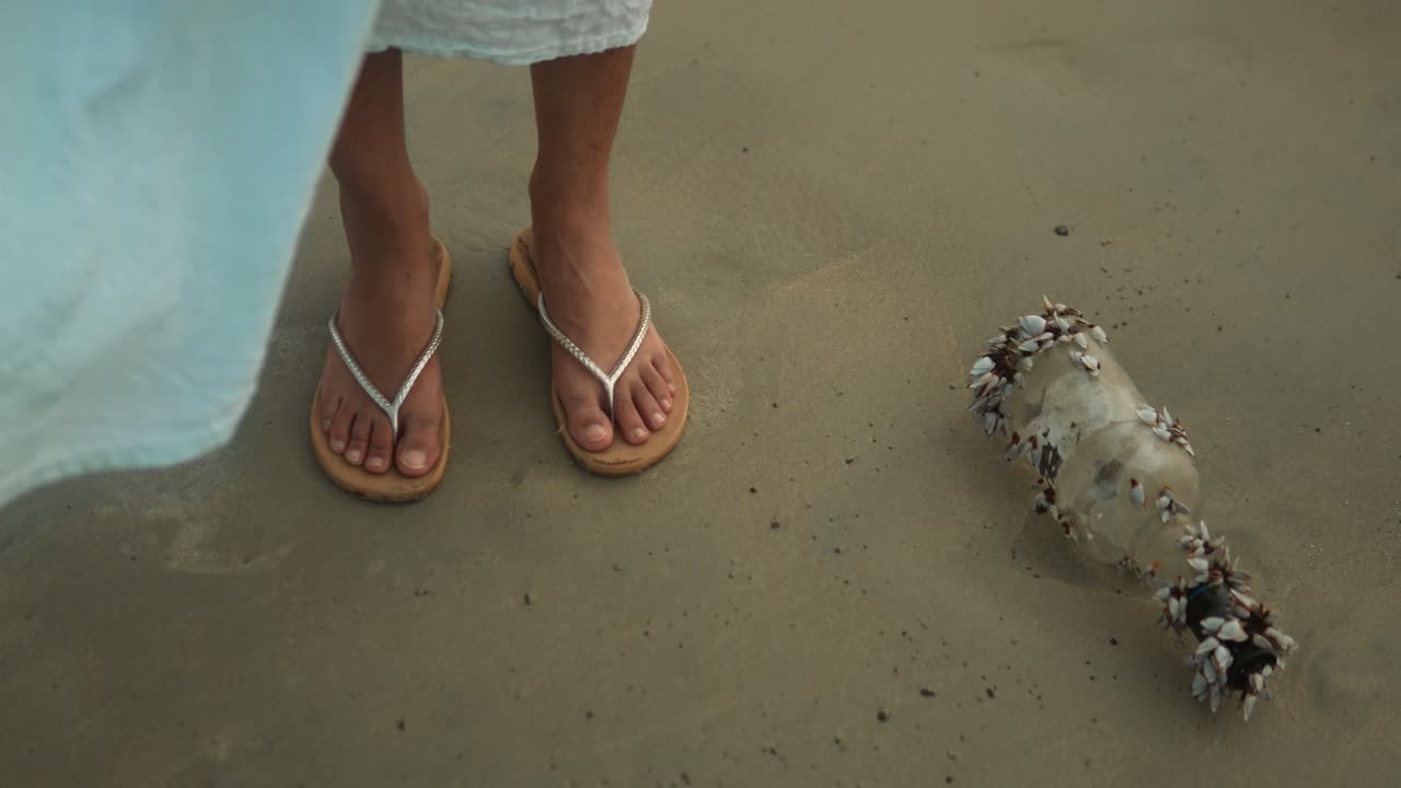 una mujer con zapatillas descubre una botella de mensajes cubierta de conchas en una playa de arena.