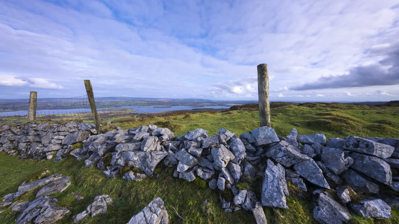 Timelapse di un paesaggio rurale con muro in pietra e pilastri in legno in un campo erboso, colline e lago in lontananza durante una giornata soleggiata e nuvolosa, visto da Carrowkeel nella contea di Sligo, in Irlanda