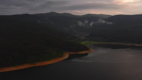 Vista-Aérea-De-Las-Montañas-Que-Rodean-Un-Hermoso-Lago-Con-Un-Pequeño-Pueblo-En-Sus-Orillas-Durante-La-Puesta-De-Sol