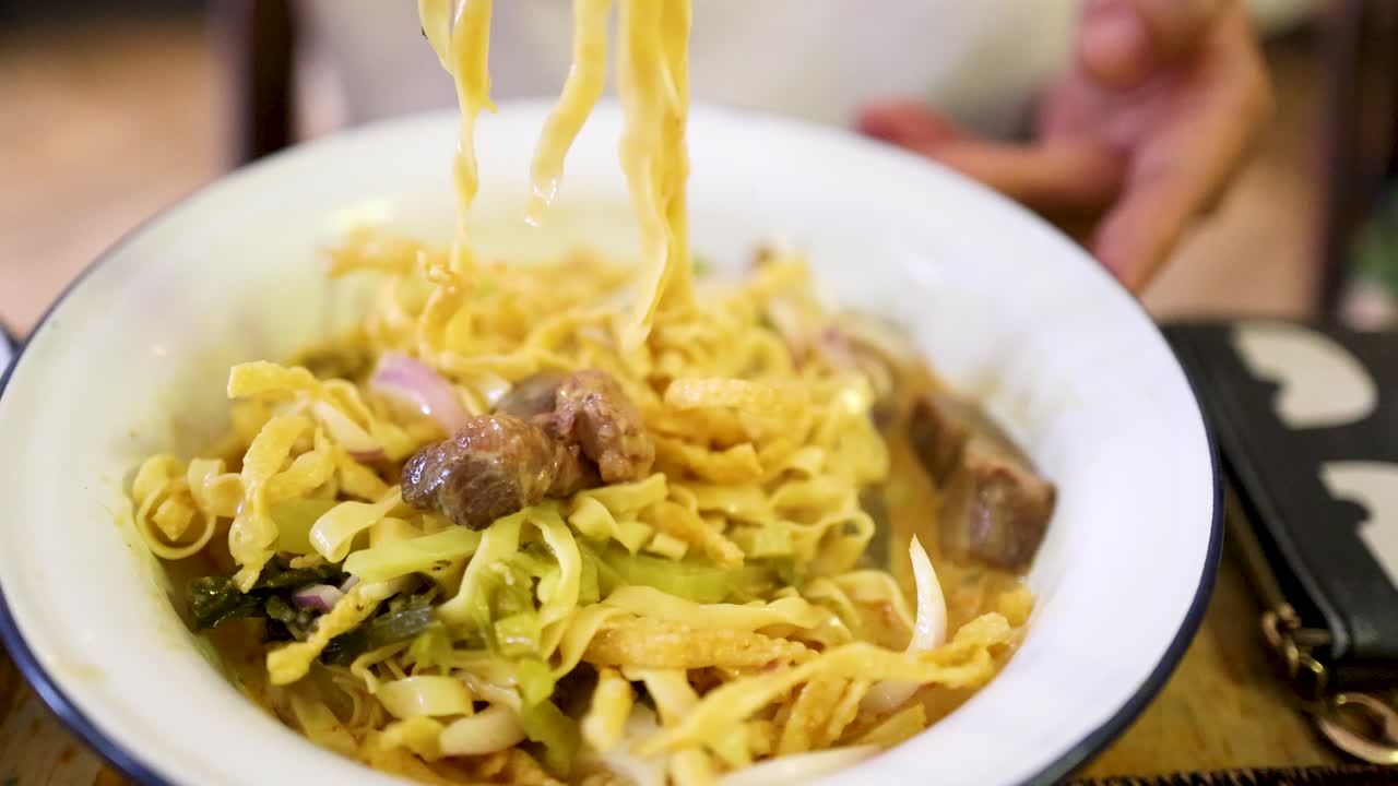 Hand uses chopsticks to mix beef curry noodles in a white bowl under warm indoor lighting, highlighting vibrant ingredients and casual dining atmosphere