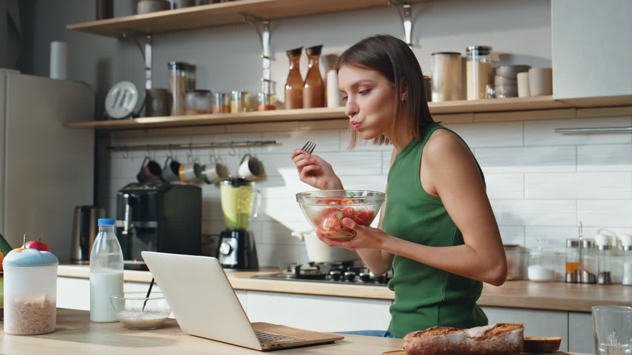 Woman eating salad while using a laptop in the kitchen