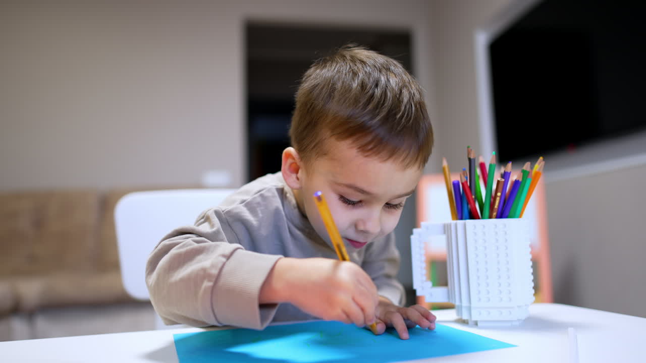 Niño pequeño disfruta de un momento de dibujo creativo. Un niño pequeño concentrado en dibujar con lápices de colores en papel azul en una mesa en casa, rodeado de materiales de arte.
