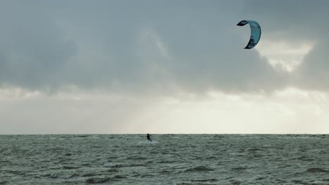 Single-kitesurfer-riding-during-storm-on-perfect-blue-water,-while-sun-is-beaming-through-dark-clouds