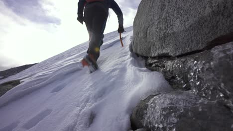 Person-hiking-up-a-snowy-hill-with-crampons-and-ice-axe,-with-partially-frozen-water-in-foreground