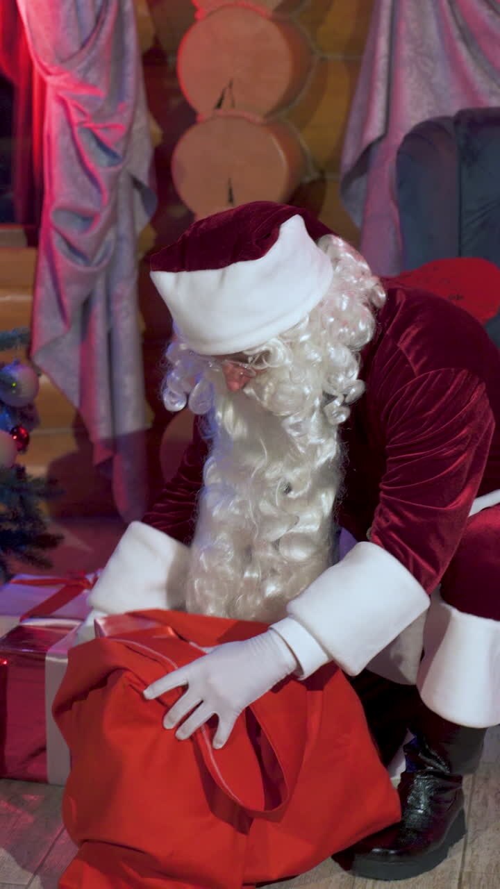 Santa Claus with a red sack indoors. Real Santa putting presents under the Christmas tree quietly. Vertical video