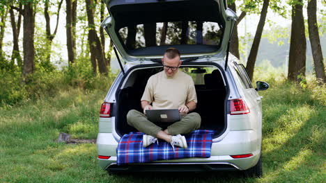 Caucasian man sitting in the trunk of his car puts on glasses. Freelancer is getting ready to work on his laptop. Beautiful summer nature at backdrop.