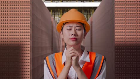 close up of asian female engineer with safety helmet pray for something while standing with red brick packed in stacks are stored