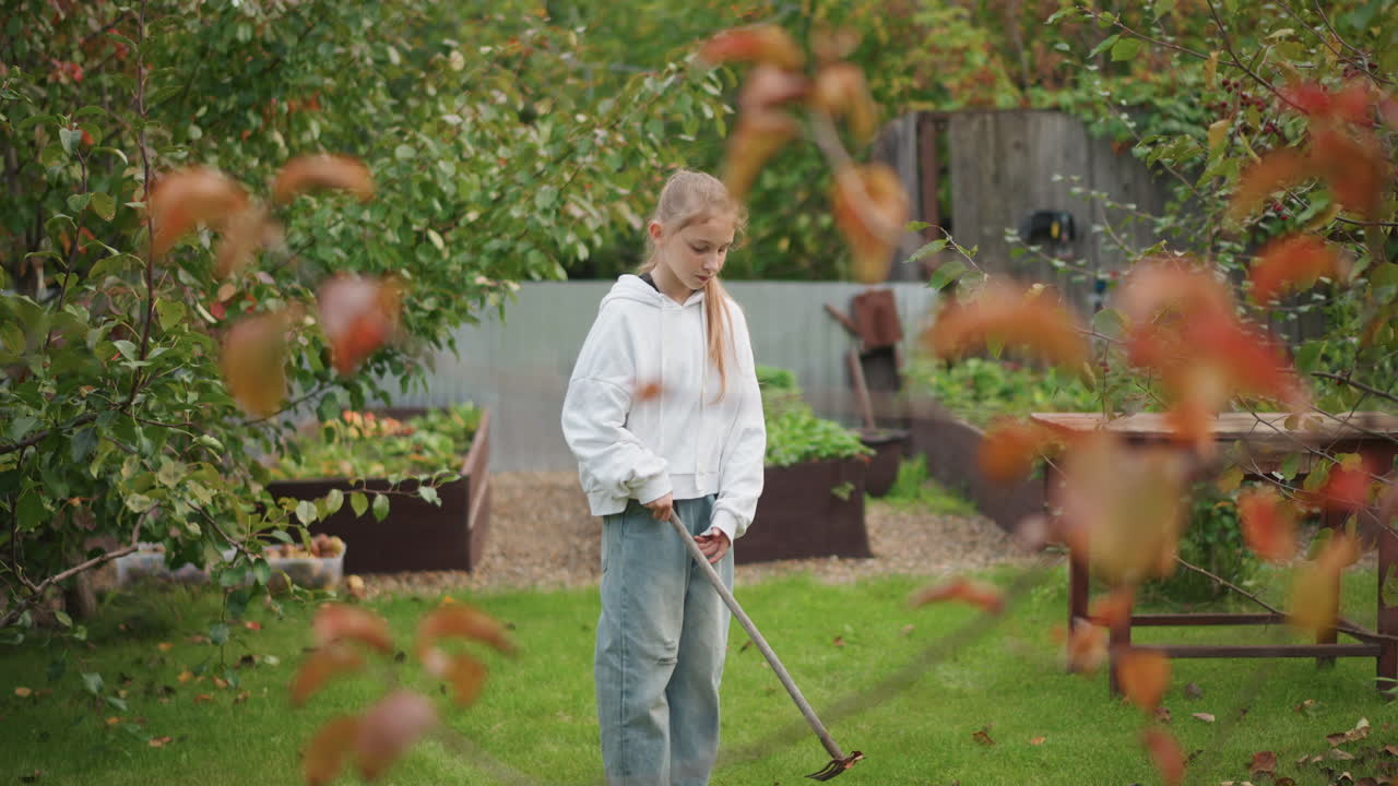 Young white girl practices garden raking near raised beds, focus on technique and routine, hands on tools, cool autumn palette with orchard plants and compost area visible, slow calm motion conveying