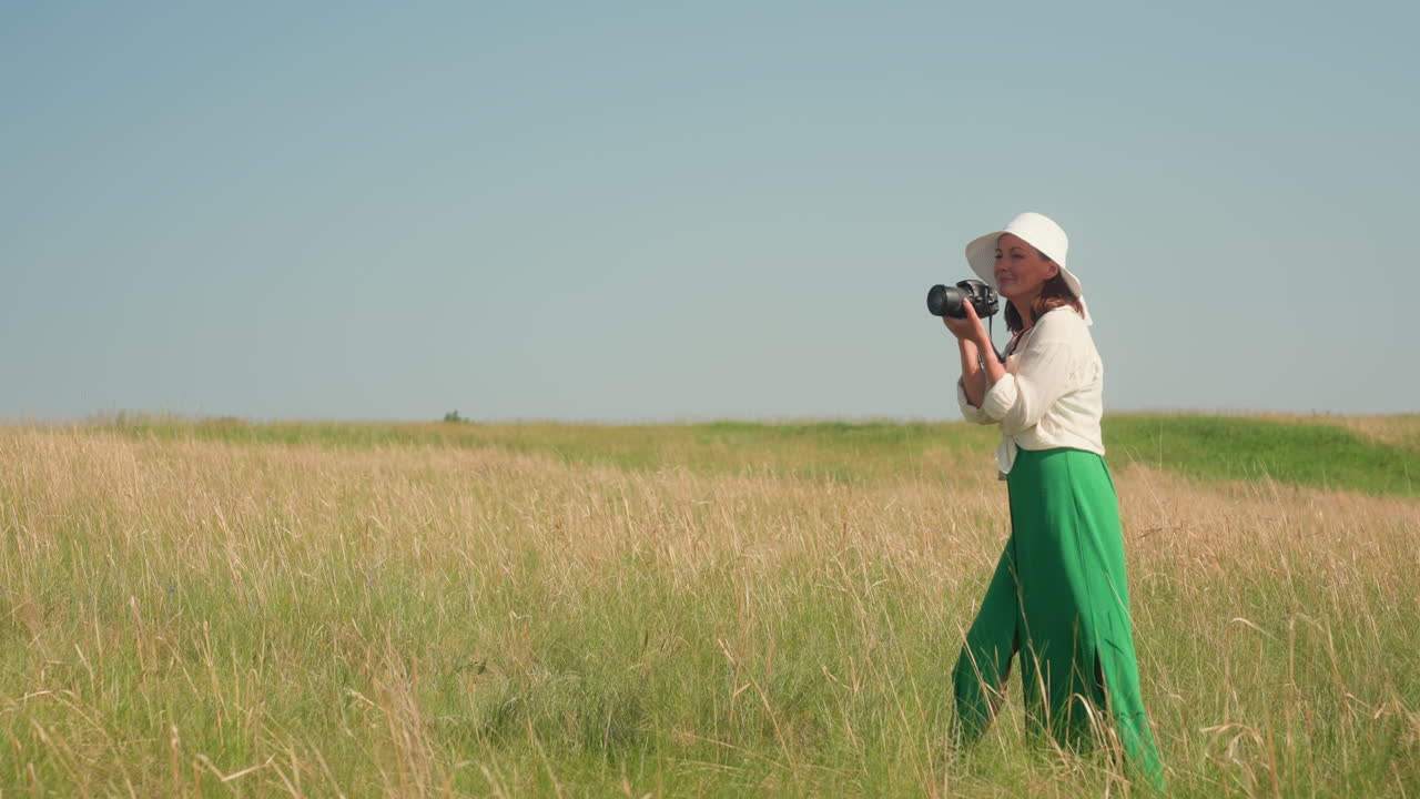Signora sorridente con cappello bianco e pantaloni verdi che tiene una macchina fotografica e scatta una foto attraverso una campagna dorata, con un campo erboso aperto e un cielo blu vivido sullo sfondo durante una luminosa giornata estiva.