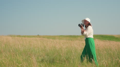 smiling lady in white hat and green trousers holding camera takes photo across golden farmland with open grassy field and vivid blue sky in background during bright summer day