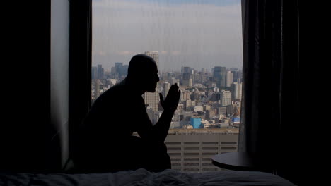 고층 아파트 창문 근처에 불안하게 앉아있는 외로운 남성의 실루, 외로움, 생각과 불안을 보여주는 뒤로 흔들리고 있습니다.