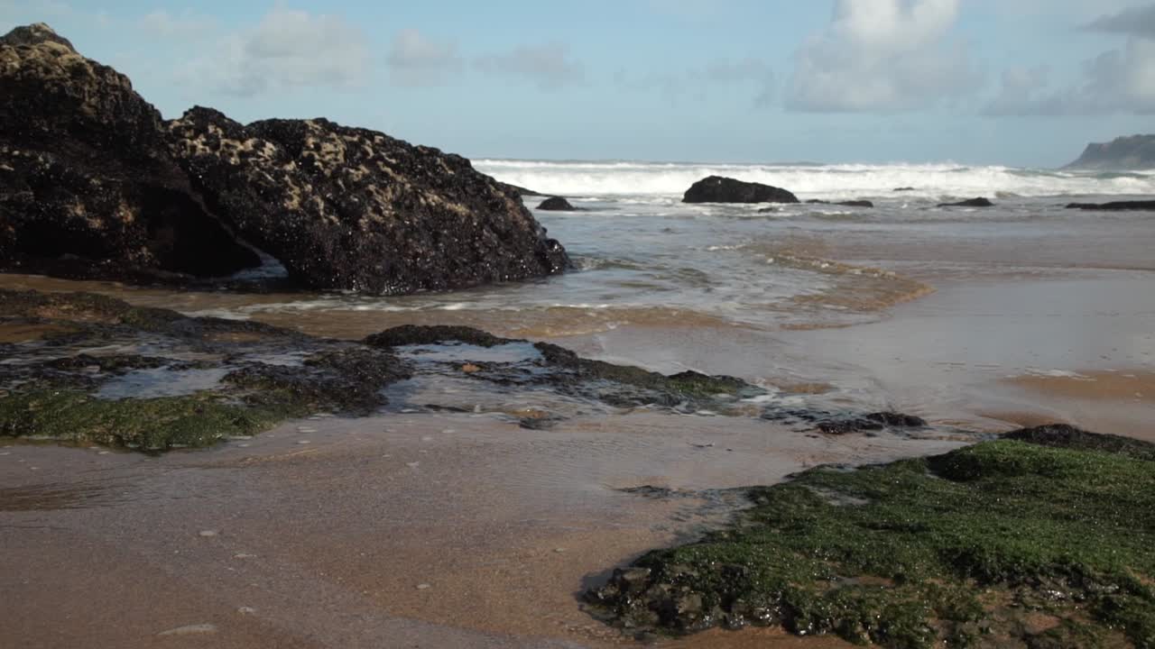 Ondas do mar inundando a praia arenosa. Rochas cobertas de algas. Praia da Cresmina, Portugal