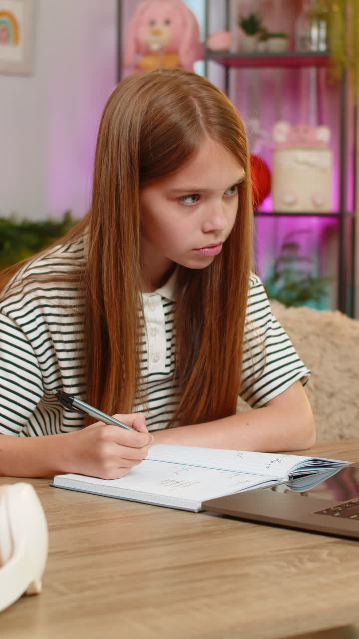 Child girl kid doing online homework on laptop and writing math notes while sitting at home table
