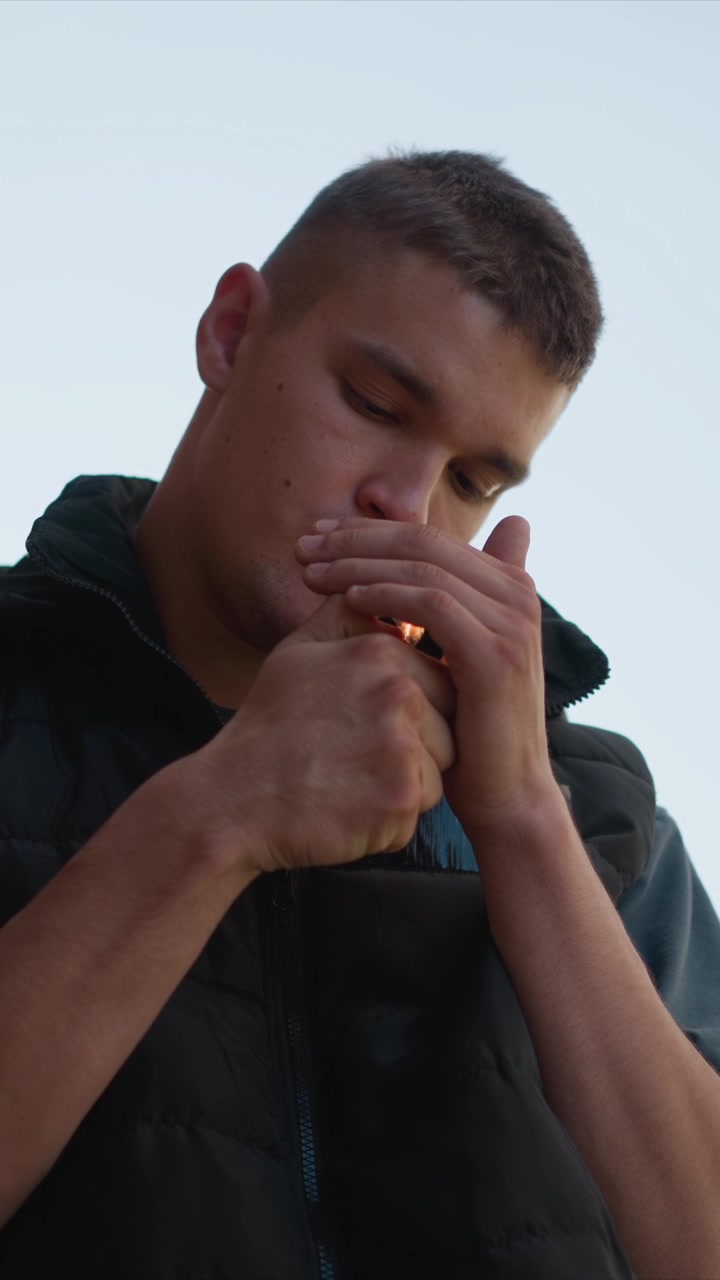 Close-up de um homem vestindo colete acolchoado preto e camisa cinza, segurando um objeto perto da orelha. O sujeito permanece praticamente imóvel, com movimento mínimo das mãos, enquanto interage com o item contra um fundo claro.