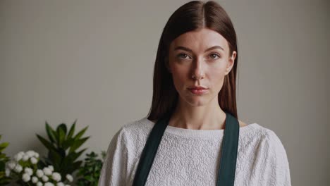 young woman florist wearing apron, with a neutral expression, looking directly at the camera, in front of a gray background and blurred plants, isolated on white