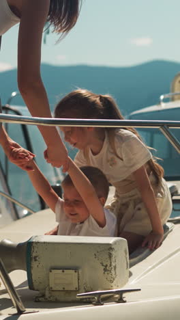 mother pulls out little boy of boat hatch with girl in sunny city port on summer day. cute children with woman leave moored yacht against mountains