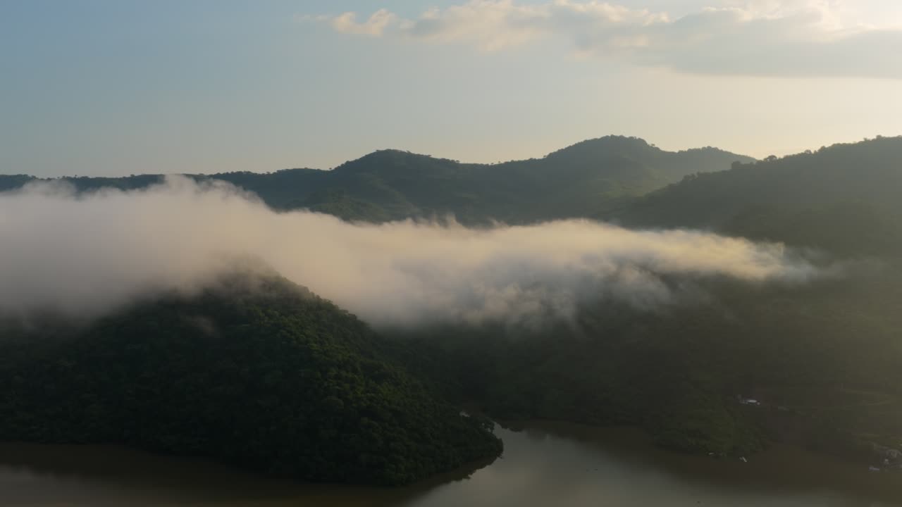 Drone video showcasing a serene, mist-covered landscape at Presa EL Carrizo, Jalisco. Golden hour light bathes the undulating hills and tranquil water below, creating a mystic atmosphere