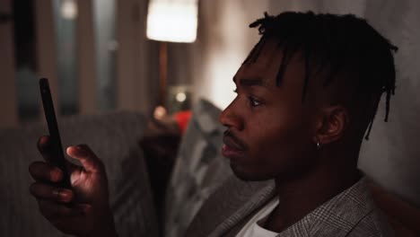 a young black male scrolls on smart phone while sat on the sofa relaxing