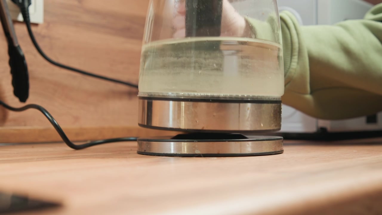 Elektrischer Glaswasserkocher wird von metallischer Basis auf warmer Holzarbeitsplatte gehoben, während Wasser blubbert und Dampf durch den transparenten Körper aufsteigt; Hand mit hellgrünem Ärmel erzeugt Aufwärtsbewegung