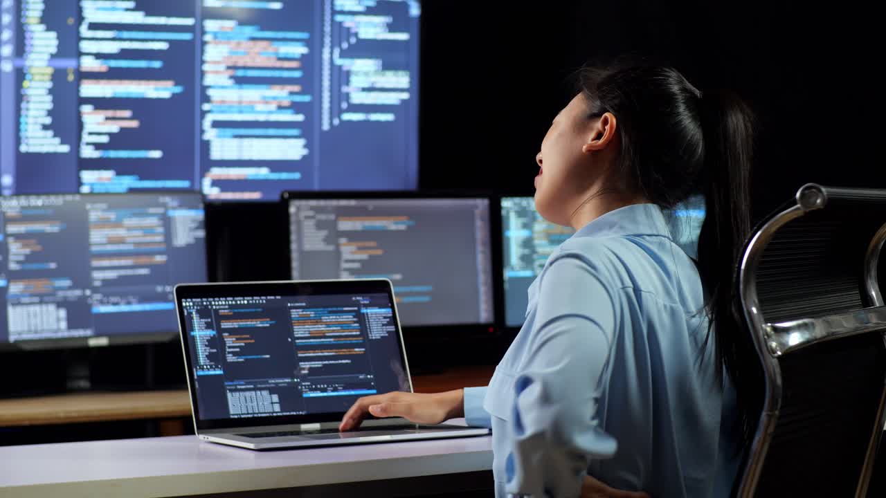 vista trasera de una programadora asiática con dolor de espalda mientras escribe código en una computadora portátil usando múltiples monitores que muestran la base de datos en los escritorios de ventana de terminal en la oficina