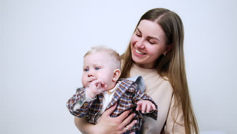 smiling long-haired caucasian woman sits on the floor holding her toddler baby. blond kid chews his finger and then starts to cry. white backdrop.