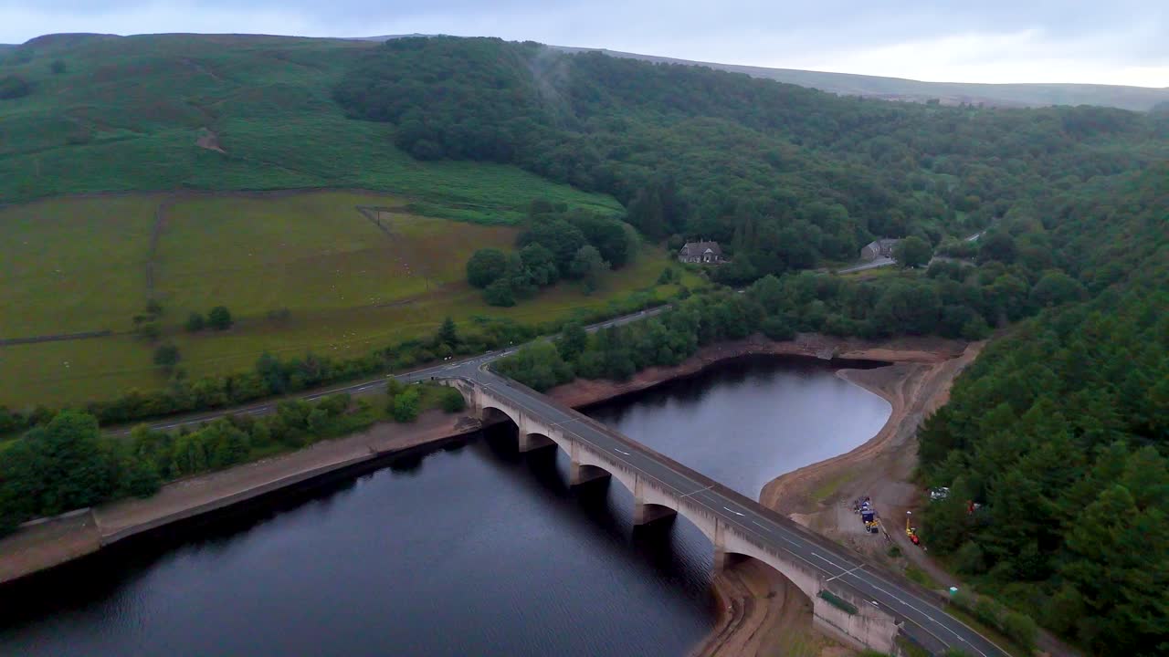 Drone footage glides above a multi-arched bridge spanning a calm reservoir, surrounded by lush green hills and forests under soft, overcast evening light