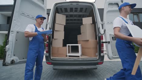 two young workers of removal company unload boxes and furniture from minibus into customer's home