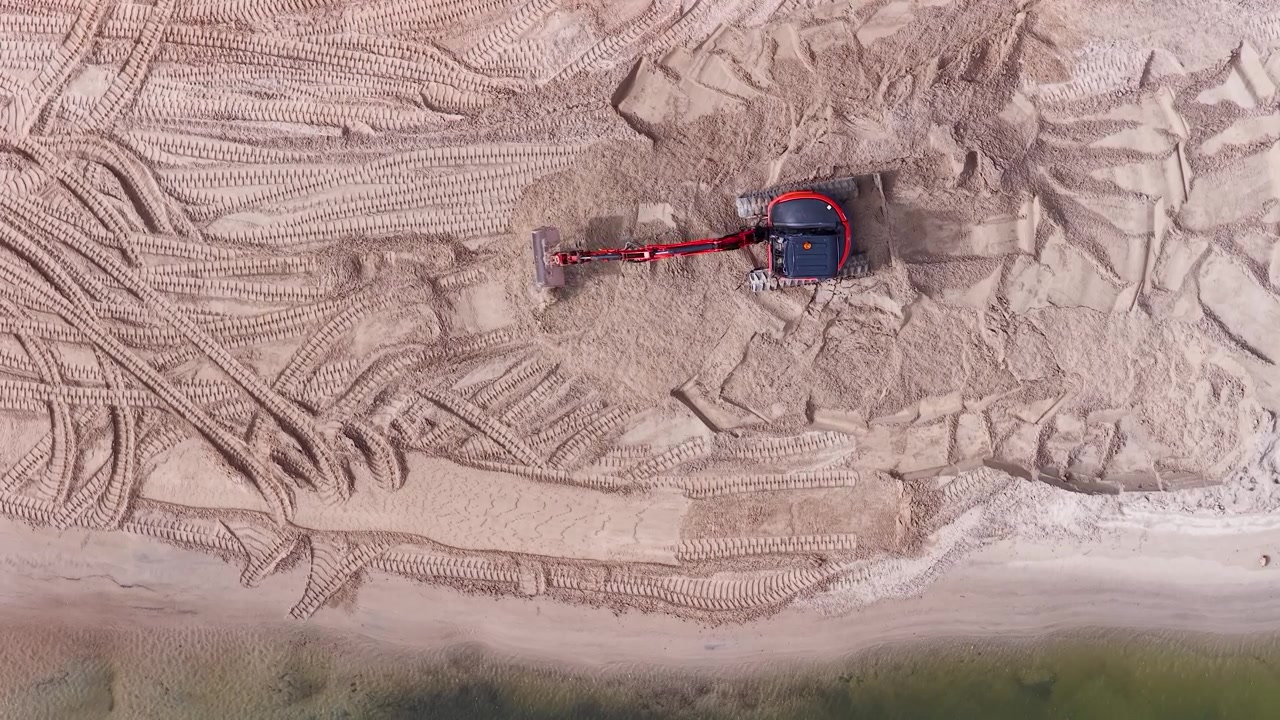 A top-down aerial shot shows a red excavator moving sand for coastal management