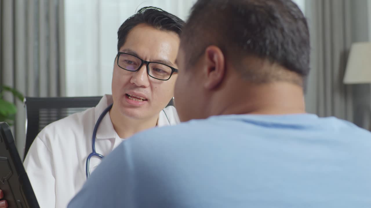 アジア人男性医師と太った男性患者が診療所でテーブルに座ってタブレットで検査の結果について議論し悪いニュースを伝え患者を励ましています - ライブドアニュース