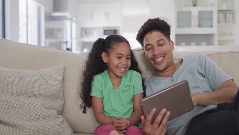 Happy-biracial-father-and-daughter-sitting-on-sofa-using-tablet